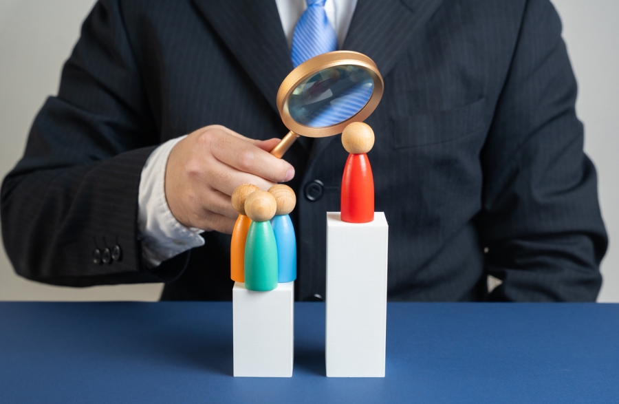 A man in a suit holds a magnifying glass over a wooden figure, illustrating the scrutiny involved in recruitment.